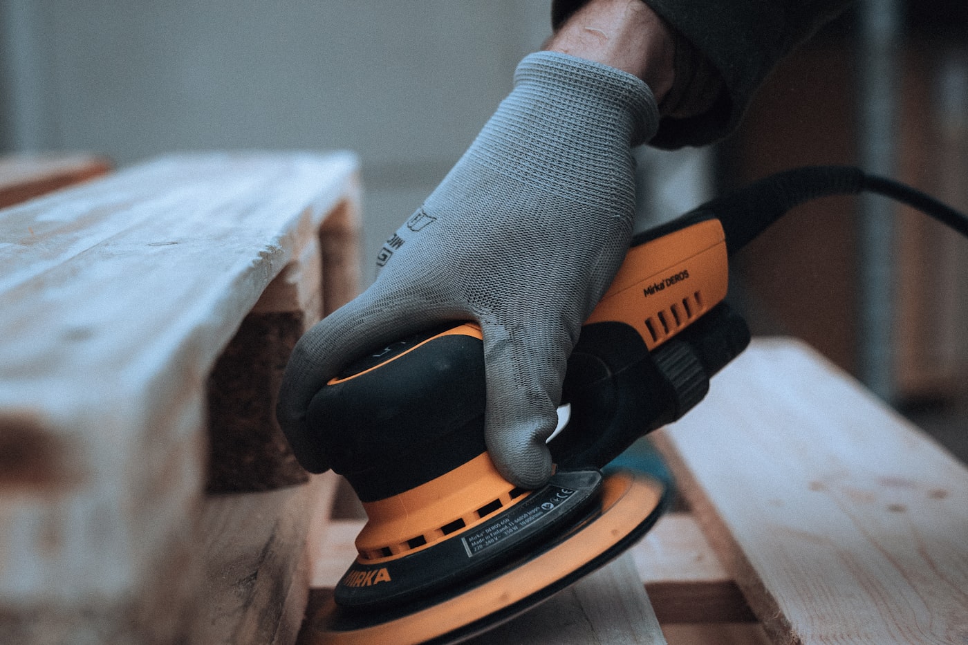 A gloved hand running a random-orbit sander along a length of dressed timber on a work stack
